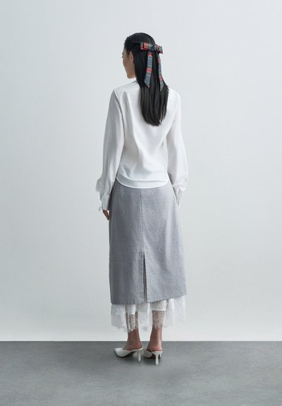 Balta garām piedurknēm blūze, kas apvienota ar pelēku linu maksi svārku ar aizmugurē esošu izgriezumu, kas kārtojusi virs baltiem mežģīņu biksēm un baltām papēžu kurpēm.