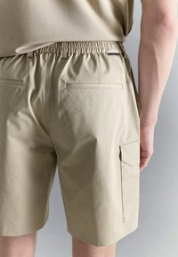 Pantalones cortos beige con cintura elástica, bolsillos de ojal en la espalda y un bolsillo cargo en el lado derecho, usados por una persona de pie frente a un fondo neutro.