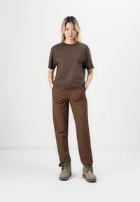 Tricou din bumbac maro cu un logo, asortat cu pantaloni maro. Modelul poartă cizme gri cu șireturi și talpă din cauciuc. Fundal neutru.
