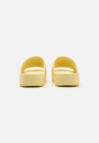 Gule glidende sandaler med tykk såle, åpen bak og avrundet design. Glatt tekstur med minimalistisk stil og horisontal mønster på sålen.