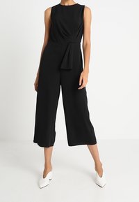 Czarny kombinezon bez rękawów z dopasowanym topem, wiązanym w pasie oraz szerokimi, skróconymi nogawkami. Gładki materiał o subtelnym połysku.