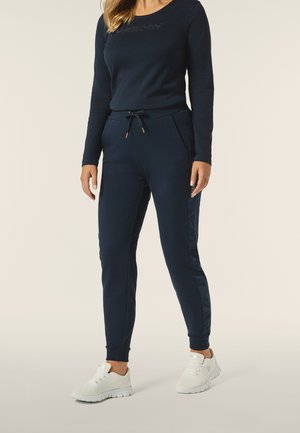 Completo da casa di colore blu navy con una maglia a maniche lunghe e pantaloni a sigaretta con tasche laterali, vita con cordino e scarpe da ginnastica bianche.