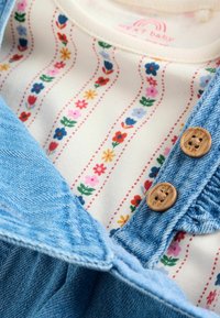 Denimruha virágmintás inggel alatta. Kék farmer anyag jellemzi, fa gombokkal és színes virágmintákkal egy krém alapon.