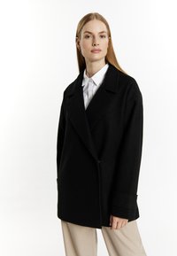 Zwarte wollen jas met een gesneden kraag, dubbele knoopsluiting en brede mouwen. Gecombineerd met een witte button-up shirt en beige broek.