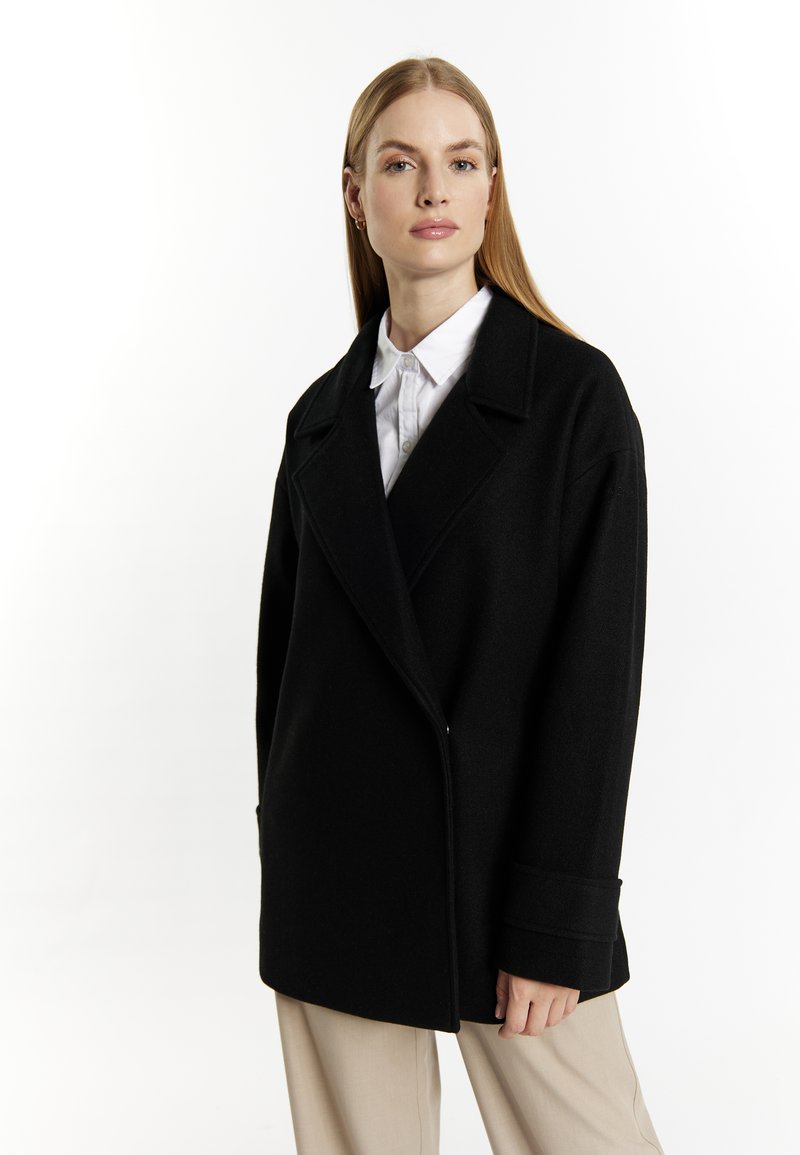 Zwarte wollen jas met een gesneden kraag, dubbele knoopsluiting en brede mouwen. Gecombineerd met een witte button-up shirt en beige broek.