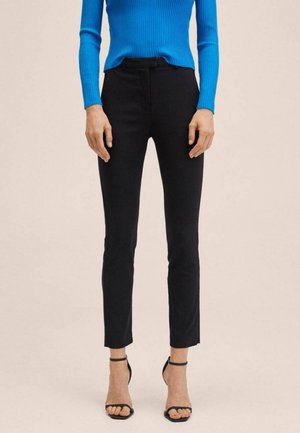 Vrouw die nauwsluitende zwarte broek, blauw shirt met lange mouwen en zwarte open-teenhakken met enkelbandje draagt, staand tegen een neutrale achtergrond.