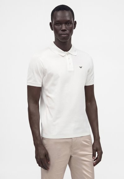 Homme portant un polo blanc à manches courtes avec un petit logo sur la poitrine, assorti à un pantalon beige, debout devant un fond uni.