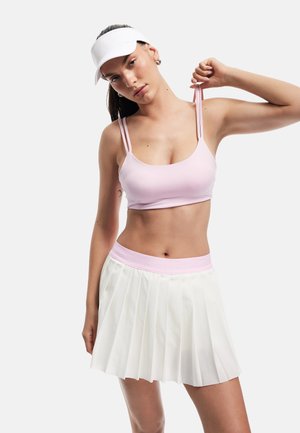 Light pink sports bra with thin straps and a white pleated skirt featuring a pink waistband. Model wearing a white visor.