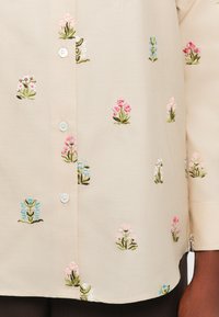 Crèmewitte shirt met meerkleurige bloemenborduursels. Het heeft een knoopsluiting met lichtblauwe knopen en een gladde textuur.