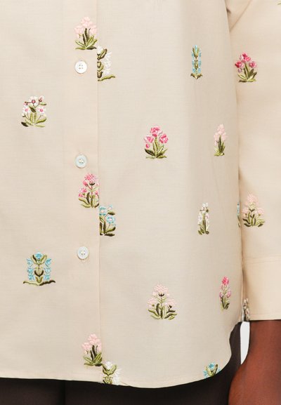 Cămașă de culoare crem, cu broderie florală multicoloră. Are un design cu închidere cu nasturi, având nasturi de culoare bleu deschis și o textură netedă.