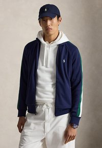 Jeune homme portant une casquette bleu marine et une veste à rayures blanches et vertes, un sweat à capuche blanc et un pantalon blanc, debout devant un fond uni.