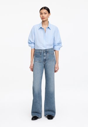 Jeune femme portant une chemise à rayures bleu clair boutonnée avec manches retroussées et un jean bleu taille haute à jambes larges avec de grandes poches avant.