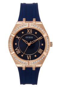 Hodinky v růžově zlatém tónu s námořnickým silikonovým páskem. Kulatý ciferník má zlaté římské číslice a bezel zdobený krystaly. Minimalistický design.