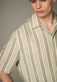 Camisa de manga corta en verde y crema con rayas verticales y una tela texturizada. Presenta un cuello y cierre de botones.