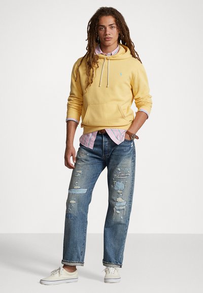 Rumena bombažna kapuca z vrvicami, plastena čez roza in belo črtasto srajco. Obuta z raztrganimi modrimi kavbojkami in belimi supergami.