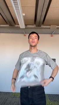 Νεαρός άνδρας σε εσωτερικό χώρο προσαρμόζει το γκρι μπλουζάκι με σχέδιο, στέκεται μπροστά σε λευκό τοίχο με βιομηχανική οροφή και στρωμένο πάτωμα με μοκέτα.