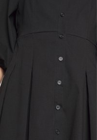 Czarna sukienka z teksturowanej tkaniny, z krótkimi rękawami, z rzędem sześciu czarnych guzików z przodu i plisowanym detalem w talii.