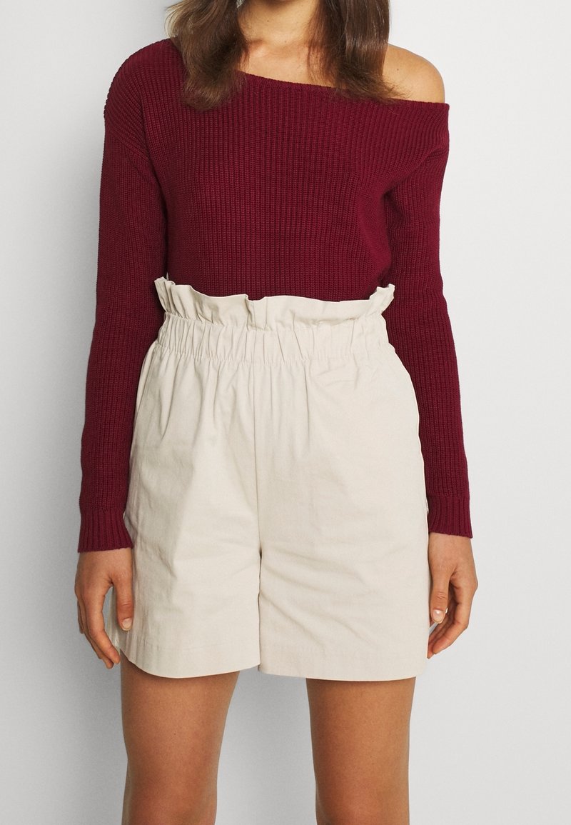 Femme portant un pull bordeaux épaules dénudées et un short paperbag beige taille haute sur un fond uni clair.