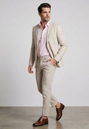 Mann, der selbstbewusst geht, trägt einen beigen Anzug, ein hellrosa Hemd und braune Lederschuhe vor einem einfarbigen grauen Hintergrund.