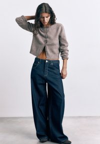 Ung kvinde iført en kort grå cardigan og mørkeblå jeans med vide ben, stående med den ene hånd bag hovedet mod en ensfarvet baggrund.