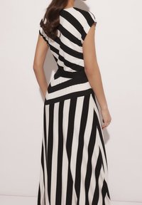 Frau, die ein schwarz-weiß gestreiftes Kleid trägt, mit diagonalen Streifen oben und vertikalen Streifen am Rock, im Vordergrund vor einem schlichten Hintergrund stehend.