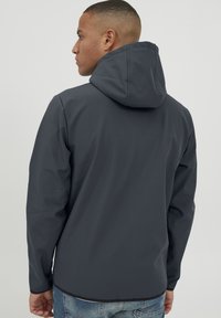 Veste �à capuche grise en matériau léger, avec un design épuré et une texture lisse, des manches longues et un ourlet se terminant juste en dessous de la taille.