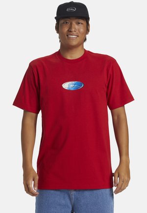 Camiseta de algodón roja con mangas cortas, con un logotipo de olas azul y blanco en el pecho. Una gorra negra está sobre la cabeza del modelo.