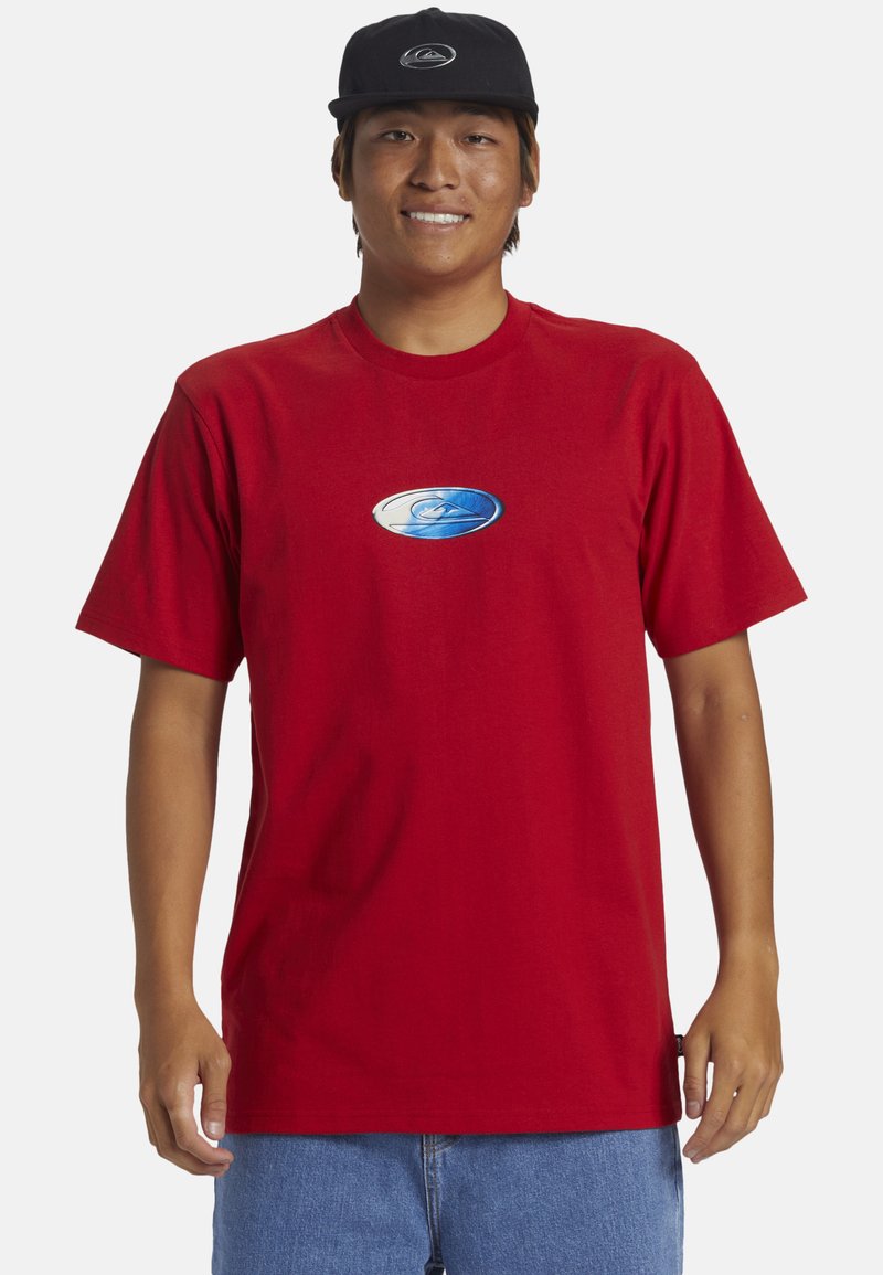 Camiseta de algodón roja con mangas cortas, con un logotipo de olas azul y blanco en el pecho. Una gorra negra está sobre la cabeza del modelo.
