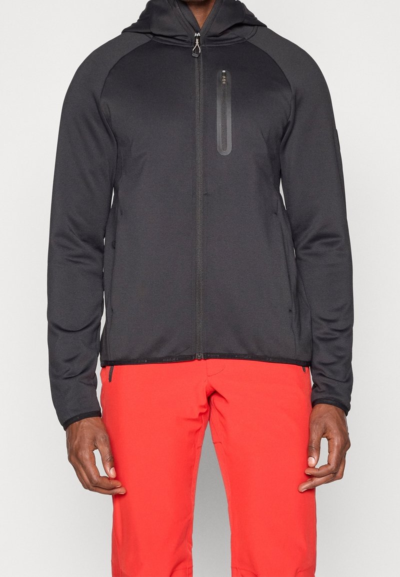 Homme portant une veste à capuche noire zippée et un pantalon rouge vif, debout avec les bras détendus le long du corps devant un fond uni.