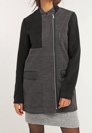 Vrouw draagt een tweekleurige grijs-zwarte jas met lange mouwen, een asymmetrische ritssluiting aan de voorkant en klepzakken, gecombineerd met een grijze rok.