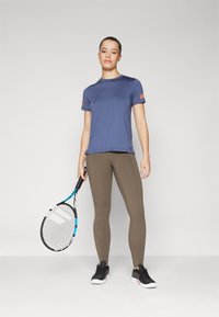 Kék haditengerészeti sportpóló rövid ujjakkal, barna leggings és fekete sportcipő. A modell egy kék és fekete teniszütőt tart.