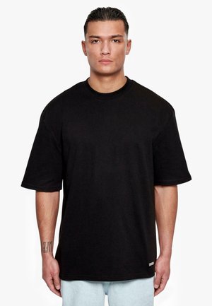 Svart oversized kortermet T-skjorte med rund hals, laget av bomull, med en subtil logotag nær sømmen og en jevn tekstur.