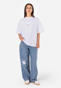 White cotton oversized t-shirt with pink embroidered text, paired with light blue baggy jeans featuring a distressed patch and white sneakers.