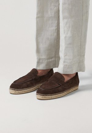 Mocasines marrones de ante sin cordones con suelas de esparto tejidas, usados con pantalones de lino de color claro.