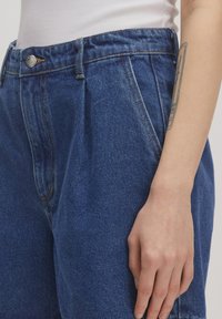Blaue Jeanshose mit hoher Taille, gefältelter Vorderseite und metallischem Knopfverschluss. Der Stoff hat eine strukturierte Oberfläche mit sichtbaren Nähten.