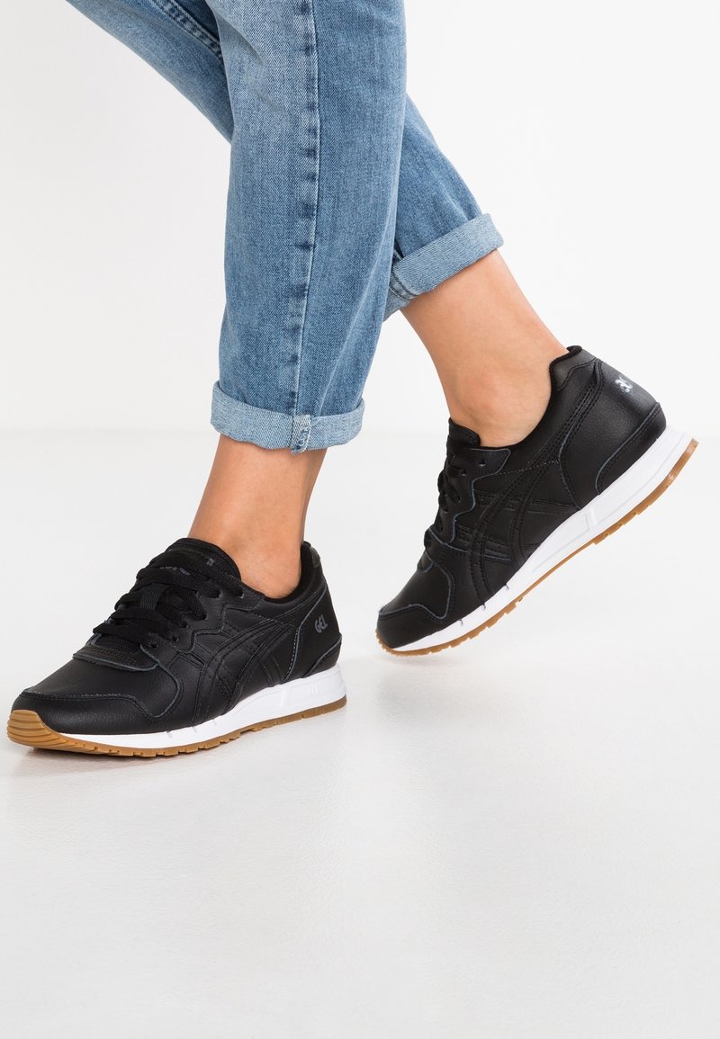 Black leather sneakers with white and gum rubber soles, textured upper, rounded toe, and black laces. Worn with cuffed blue jeans.