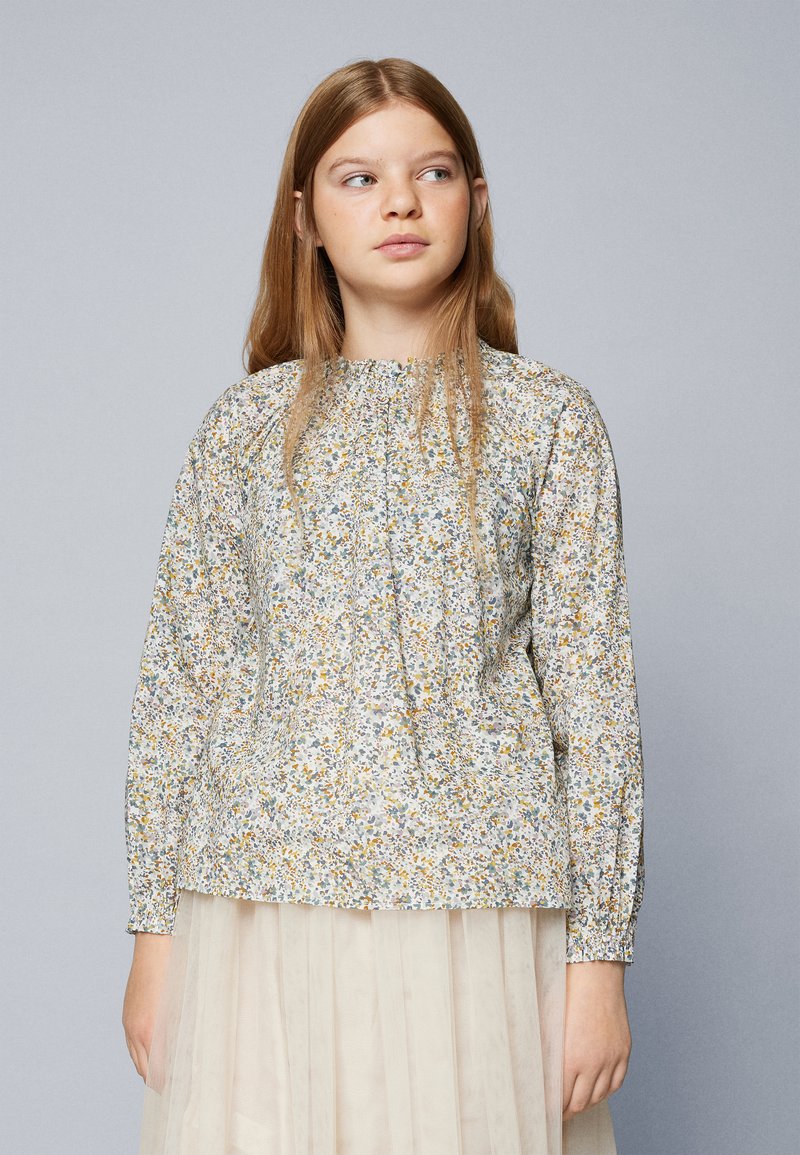 Blusa a maniche lunghe con motivo floreale in tessuto leggero, presenta un colletto arricciato e polsini elastici, con colori che includono giallo, verde e lavanda.