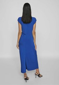 Niebieska sukienka do kolan z krótkimi rękawami, gładka tekstura, prosta sylwetka, rozcięcie z boku, noszona z czarnymi sandałami na wysokim obcasie.