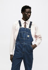 Hombre con camisa rosa claro y corbata con estampado bajo un peto vaquero azul, de pie con una mano en el bolsillo contra un fondo blanco.