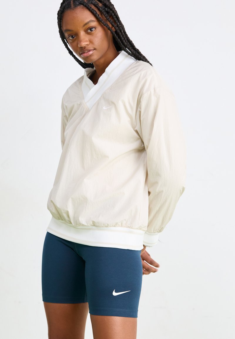 Beżowa, lekka kurtka typu pulower z białym kołnierzykiem i elastycznym dołem, zestawiona z ciemnoniebieskimi dopasowanymi szortami z białym logo.