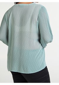 Blusa azzurro chiaro in tessuto trasparente, con un motivo a pois e maniche plissettate texture. Orlo arrotondato, design leggero e fluido.