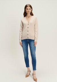 Žena v béžovom button-up svetri, modrých úzkych džínsach a hnedých lodičkách s ostrou špičkou stojí na jednolitom pozadí.