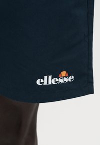 Κοντινή λήψη σκούρων μπλε σορτς με λευκό και πορτοκαλί λογότυπο ellesse κοντά στο κάτω μέρος, φορεμένα από άτομο με σκούρο δέρμα.