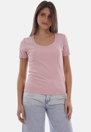 Donna con capelli castano lisci che indossa una maglietta a maniche corte rosa chiaro e jeans a vita alta azzurri con dettagli di strass.