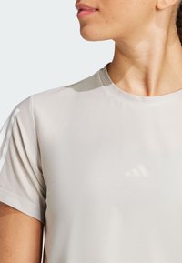 Camiseta deportiva de color gris claro con una tela texturada, cuello redondo y mangas cortas. Presenta tres franjas blancas en la manga izquierda.