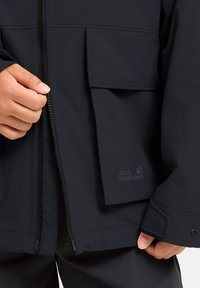 Giacca impermeabile nera con cerniera frontale, ampia tasca e dettaglio con logo. Texture liscia e design funzionale per l'uso all'aperto.