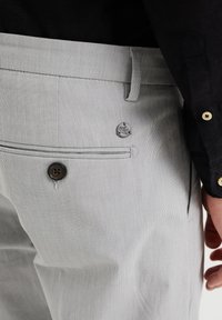 Světle šedé kalhoty vyrobené z texturované látky, s knoflíkovým zapínáním, drobným logem jako akcentem a boční kapsou pro praktičnost.