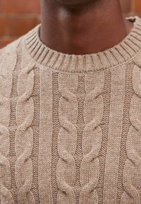 Pull en maille beige clair présentant un motif épais en torsades, un col côtelé et un tissu texturé. Motifs remarquables en rangées verticales.