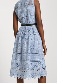 Ärmlös ljusblå spetsklänning med intrikata geometriska och blommiga mönster, åtsittande i midjan med ett svart bälte, sedd bakifrån.