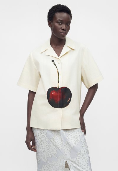 Γυναίκα που φοράει ένα κρεμ πουκάμισο με κοντά μανίκια και μεγάλο κόκκινο γραφικό κεράσι και μια λευκή υφασμάτινη φούστα με σκίσιμο εμπρός.
